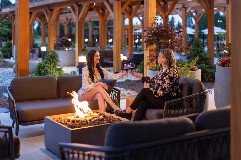 2 women sitting next to a fire drinking wine on a patio